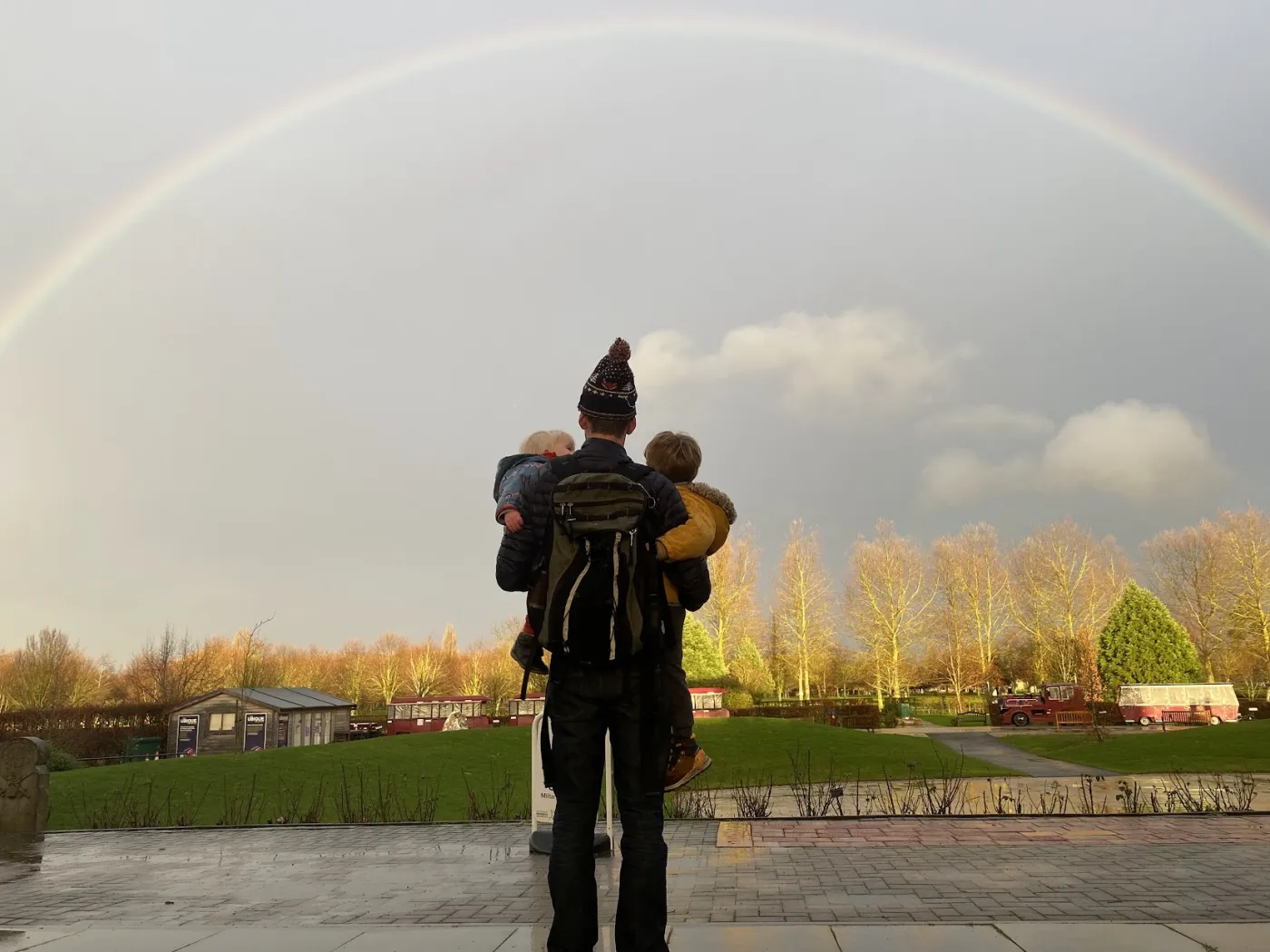 Nick, seen from behind, carrying his two young children towards a full rainbow over a park.